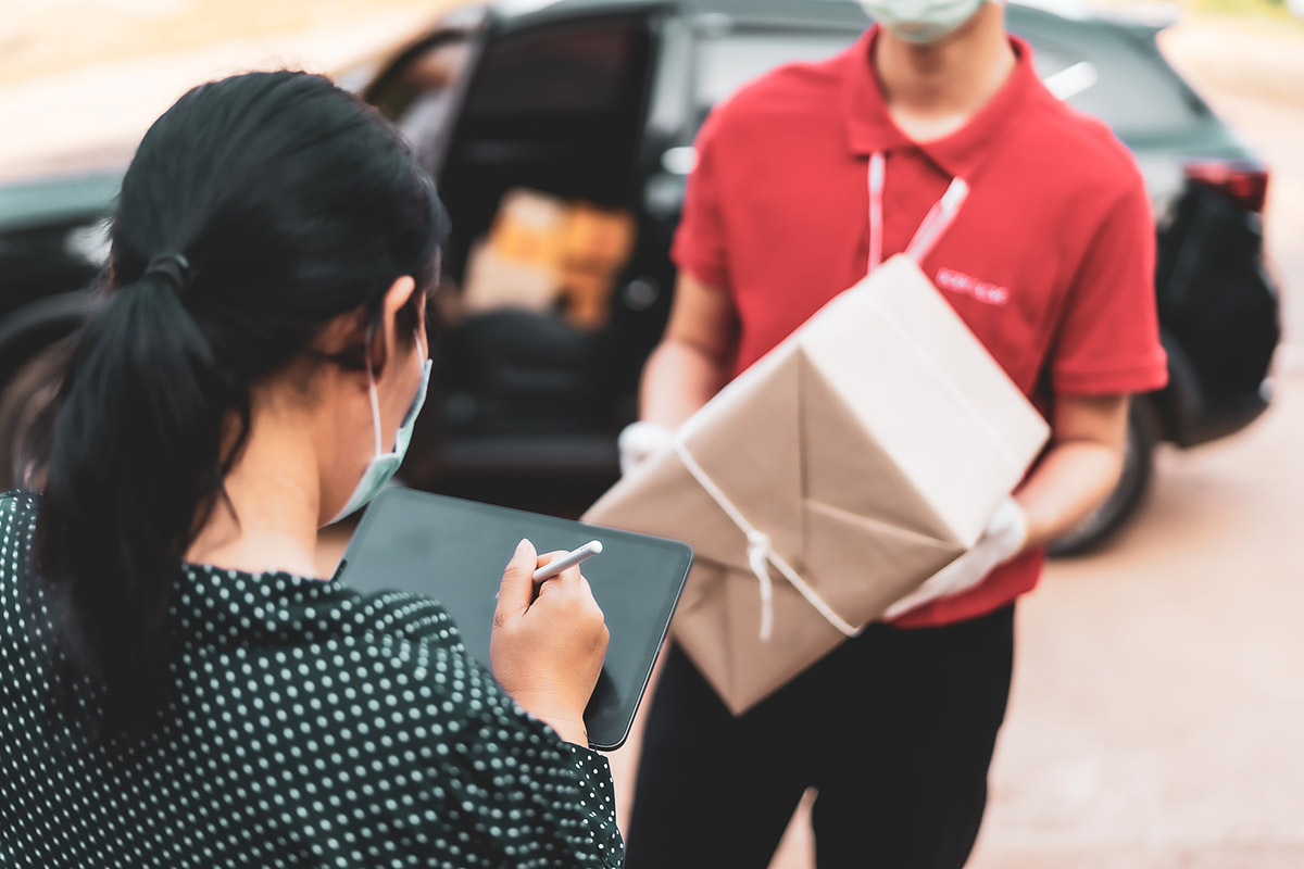 Bud levererar paket till kvinna som signerar leveransen på en pekskärm. I bakgrund syns en mörk personbil med fler paket i.