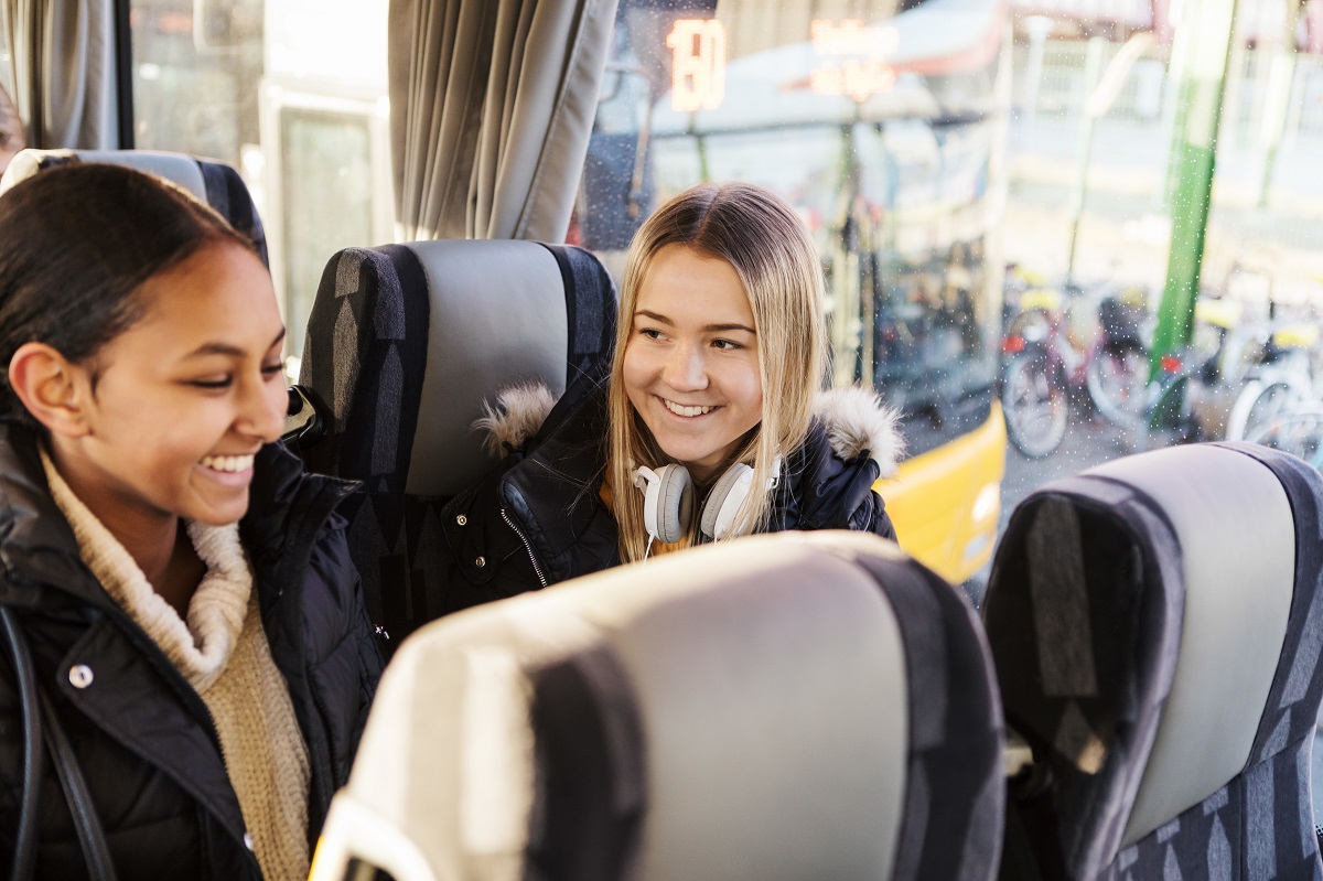 Två glada ungdomar sitter i en buss. I förgrunden finns fler bussäten. Utanför bussfönstret finns ytterligare en buss och många parkerade cyklar.